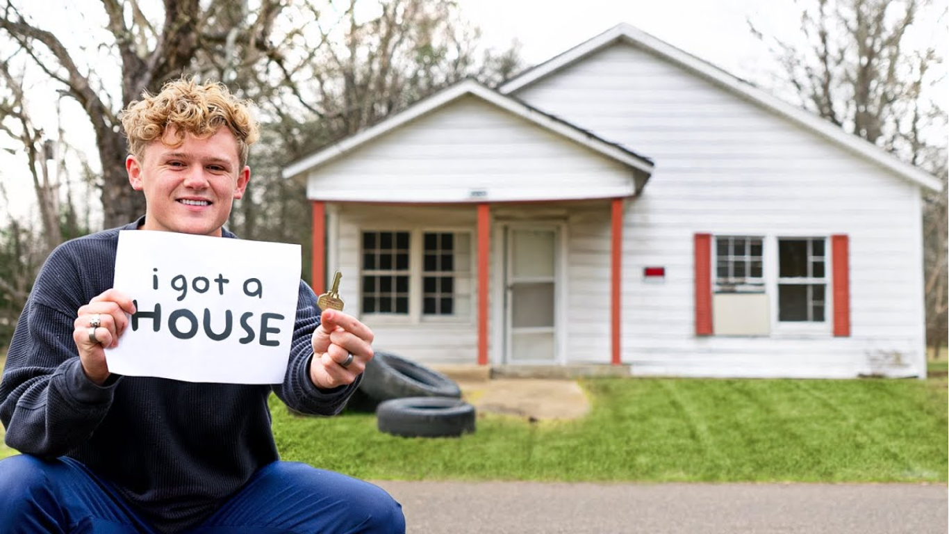 Ryan Trahan sitting in front of a house holding a key and white cardboard